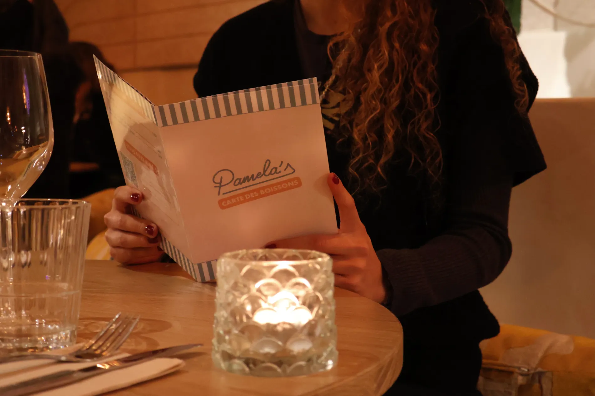 Close-up of a customer holding the retro drinks menu at Pamela's Bar in Nice, seated at a wooden table lit by a candle holder.
