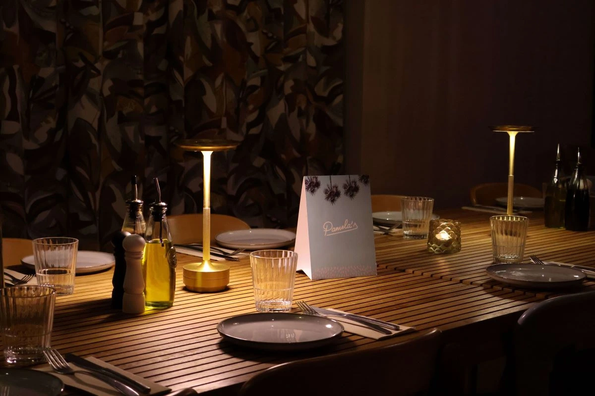 Long wooden table set for dinner and illuminated by golden cordless lamps in a cozy, dim-lit room at Pamela's Bar in Nice.